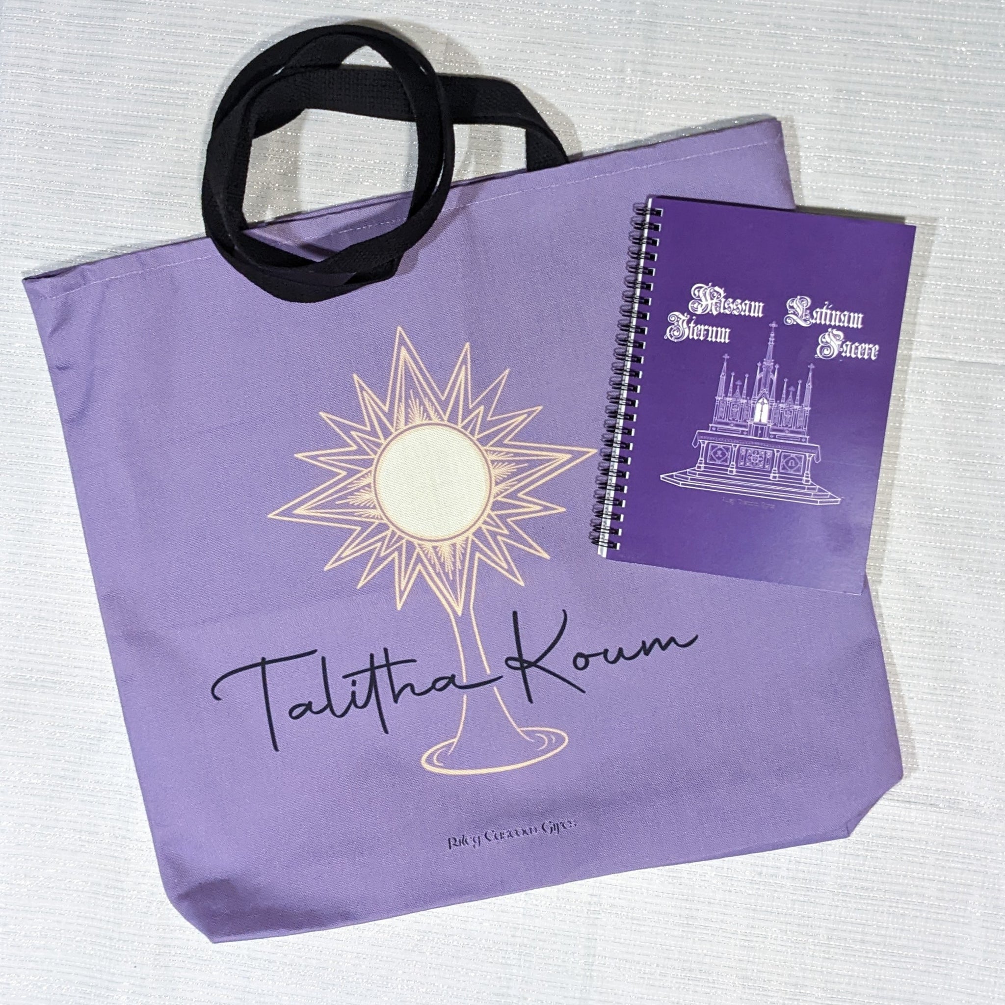 A Catholic tote bag and Catholic notebook, both purple. The tote bag has a monstrance and Talitha Koum on it, and Make Mass Latin Again design on the notebook