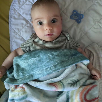 A baby looks up at the camera with a Catholic blanket over it featuring rainbows, doves, and olive branches representing Noah's Ark