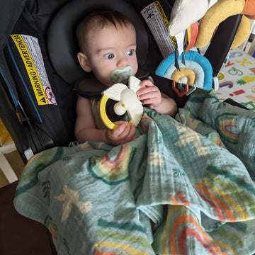 A baby in a car seat with a Catholic swaddle featuring rainbows and doves to represent Noah's Ark