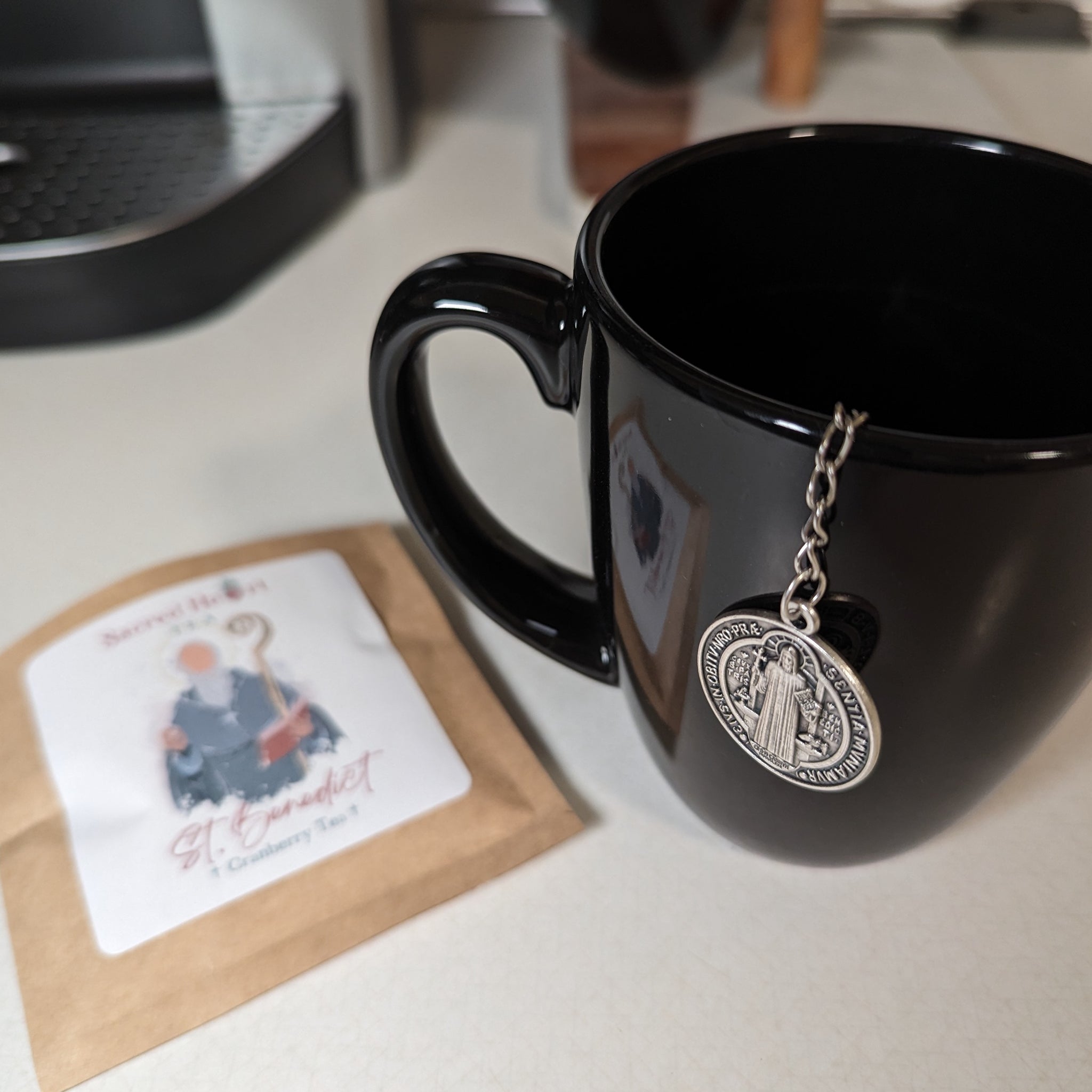 A black mug with a Catholic tea infuser featuring the St. Benedict medal next to St. Benedict loose leaf tea