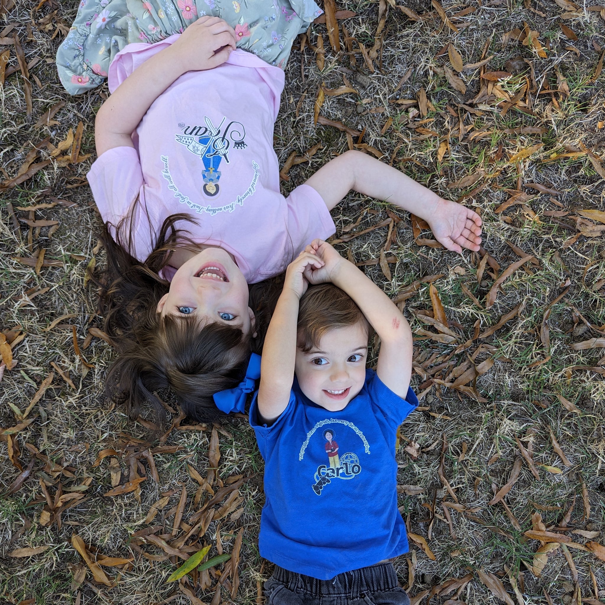 A boy and girl lying on the ground looking up at the camera. The boy is wearing a Bl. Carlo Acutis blue shirt and the girl is wearing a pink St. Joan of Arc shirt.