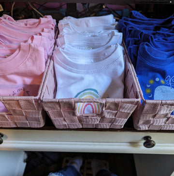 Three baskets of Catholic toddler t-shirts in pink, white, and blue with Catholic symbols on them