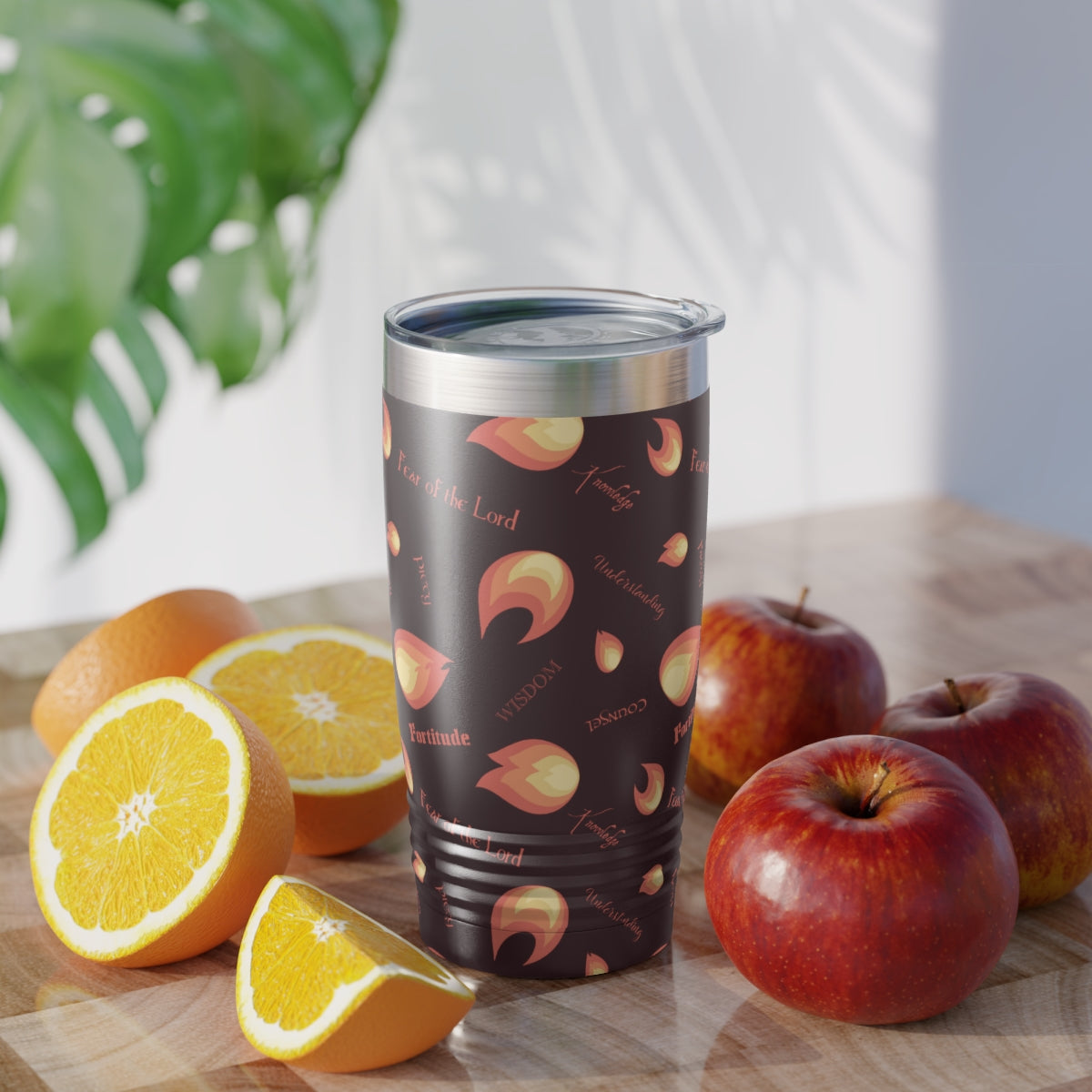 A brown Catholic travel mug on a table with apples and oranges. The mug is decorated with flames and the gifts of the Holy Spirit written out.