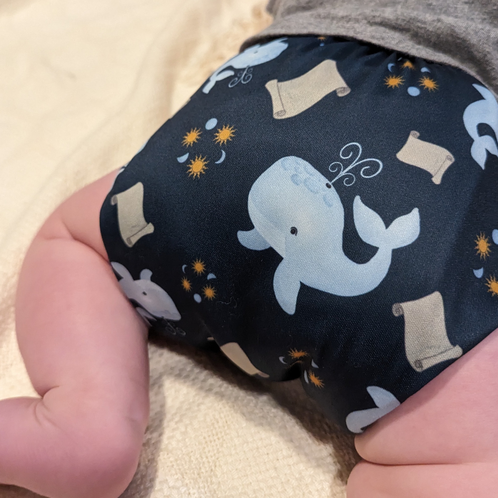 A Catholic cloth diaper on a baby featuring the Jonah's Wait design with whales, scrolls, and three suns and moons