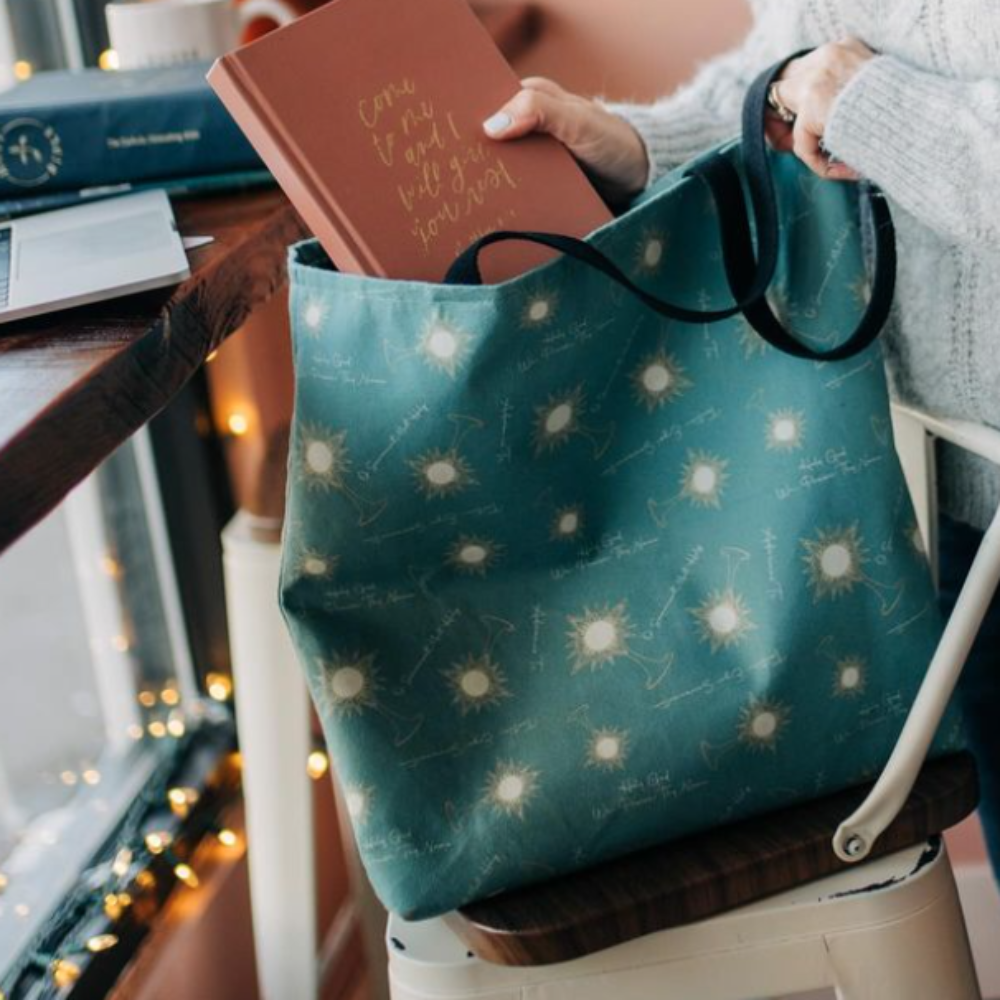 A woman putting a devotional into a Catholic tote bag sitting on a chair. The tote bag is teal with monstrances and the words from Eucharistic Adoration on it.