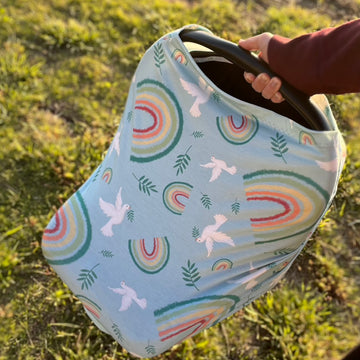 A woman's arm holding a car seat with a Catholic car seat cover featuring rainbows, doves, and olive branches