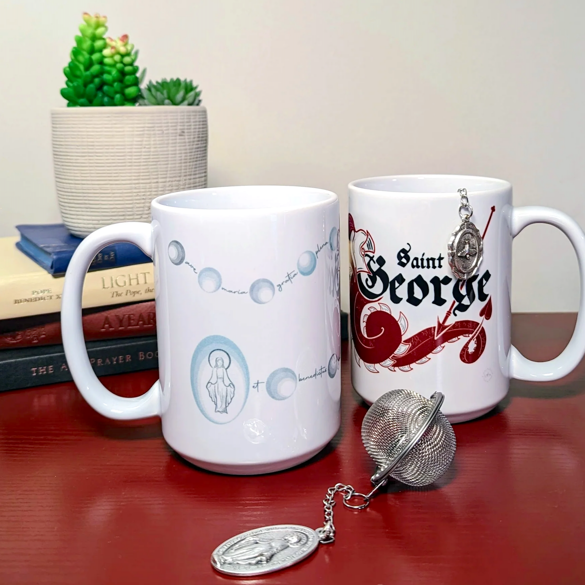 Two Catholic mugs. One mug has a rosary and miraculous medal design while the other mug has a dragon and the name "Saint George" on it. A miraculous medal tea infuser is displayed in front of the two mugs. A Holy Spirit medal is displayed off the side of the St. George mug.