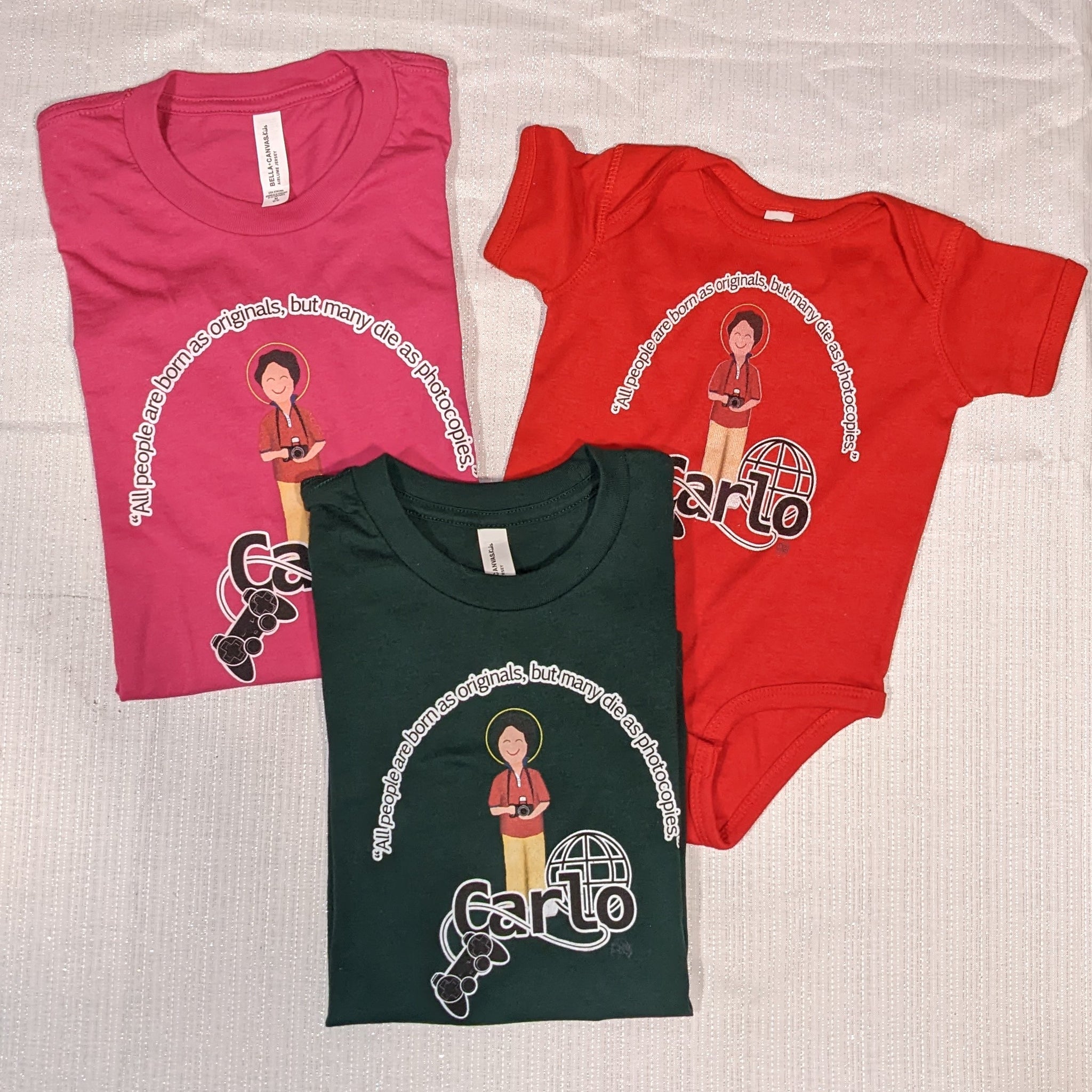 Three Catholic kids shirts, one a baby onesie, featuring Bl. Carlo Acutis and the phrase "All people are born as originals but many die as photocopies."