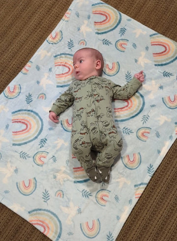 Baby sitting on a Catholic blanket with a design that features a rainbow, dove and olive leaf design to represent "God's Promise".