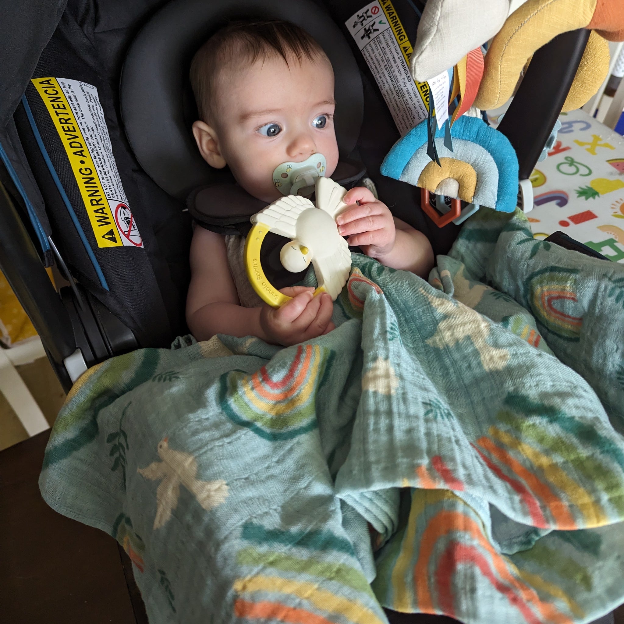 A baby in a car seat with a Catholic swaddle blanket with rainbows, doves, and olive branches. The baby is holding a Holy Spirit teether.