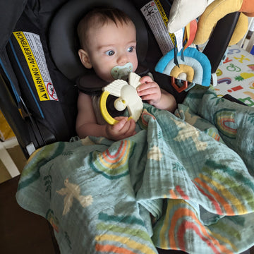 A baby in a car seat with a Catholic swaddle blanket with rainbows, doves, and olive branches. The baby is holding a Holy Spirit teether.