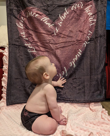 Baby sitting in front of a Catholic blanket while wearing a Catholic cloth diaper. Both the diaper and the blanket feature the St. Therese of Lisieux quote "Let us love since that is what our hearts were made for."