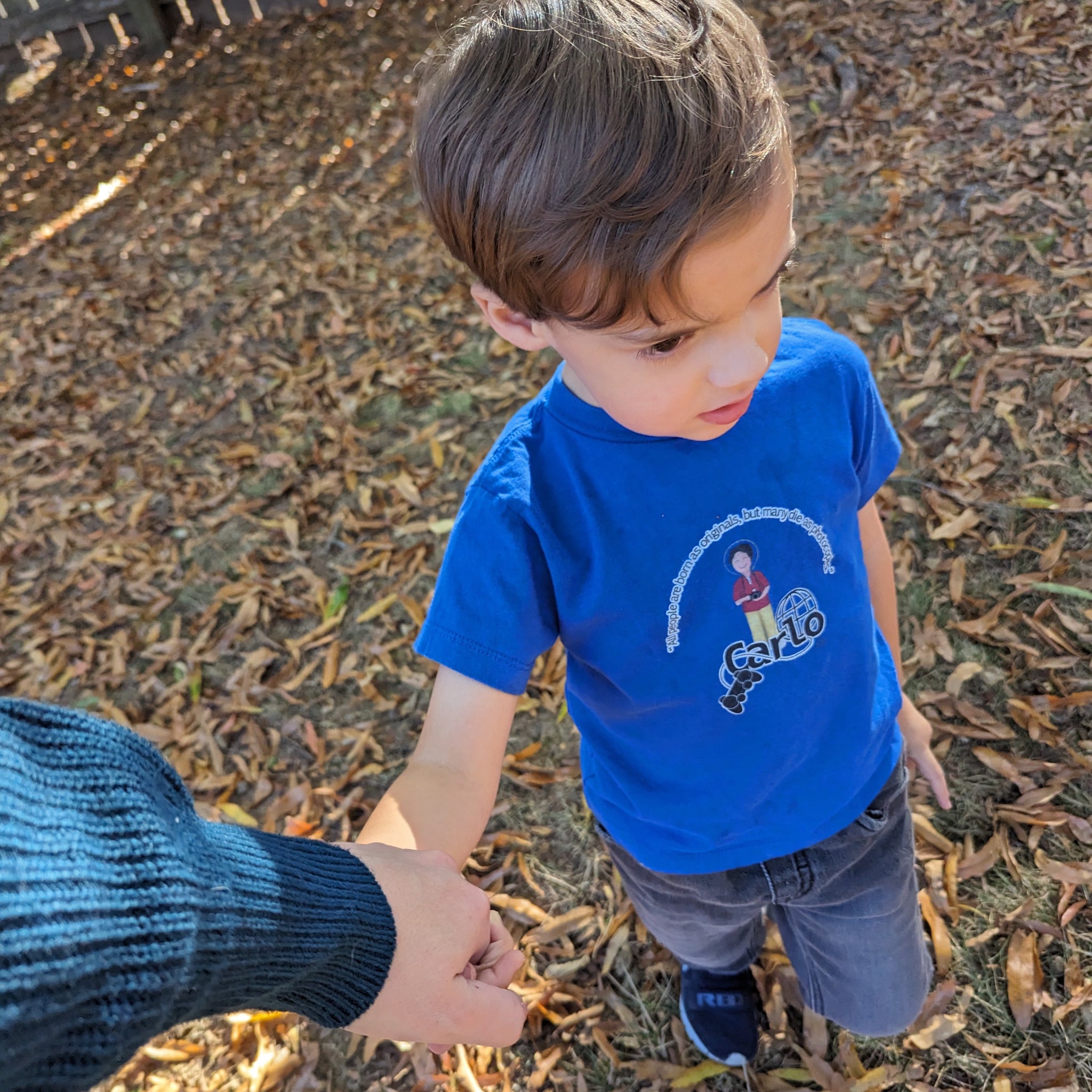 Kid wearing a blue shirt featuring Bl. Carlo Acutis and the quote "All people are born as originals, but many people die as photocopies."