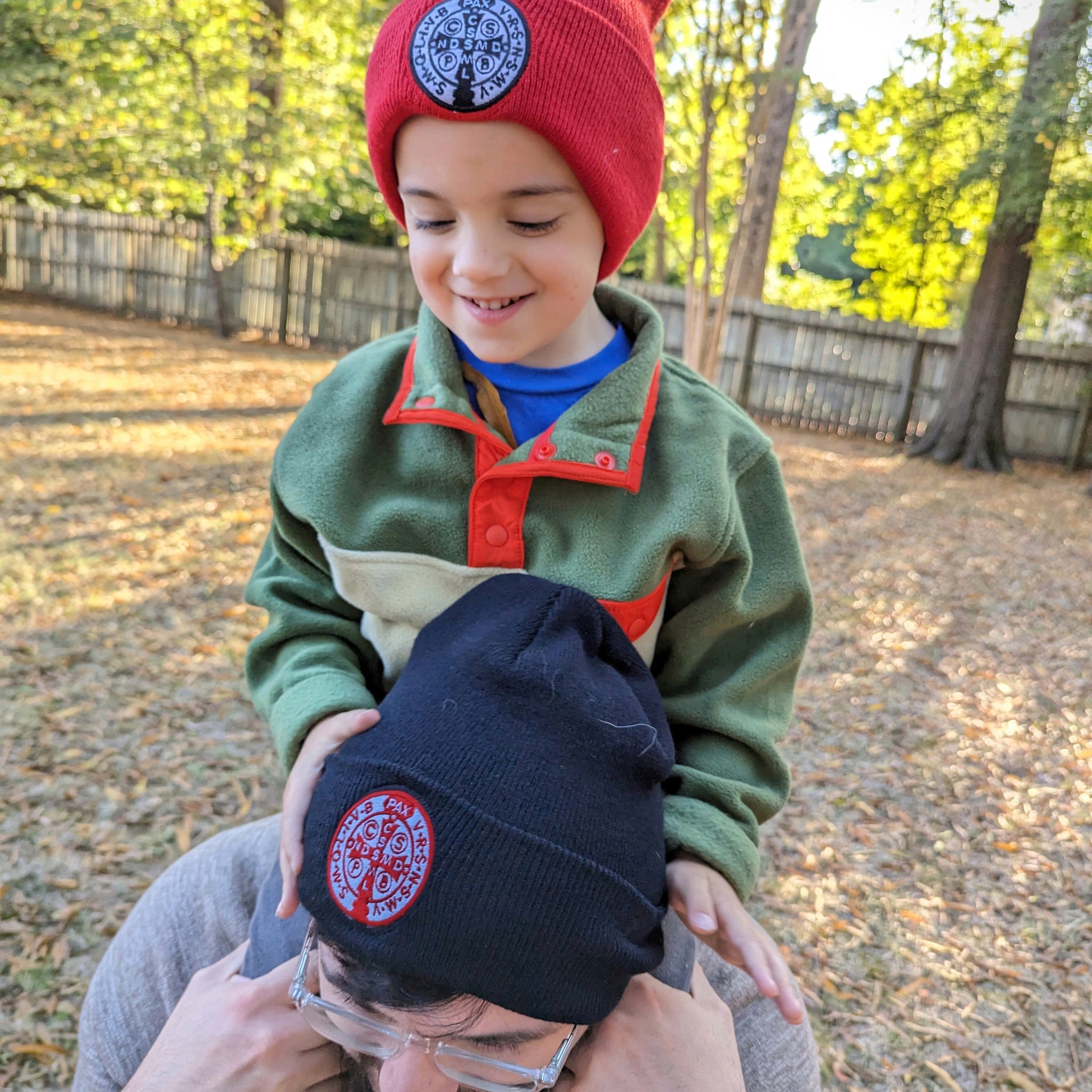 Kid sitting on man's shoulders. Both the boy and the man are wearing Catholic beanies, or hats, with the St. Benedict medal stitched onto the hats.