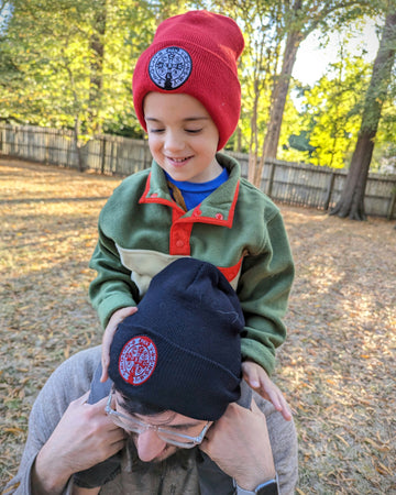 Kid sitting on man's shoulders. Both the boy and the man are wearing Catholic beanies, or hats, with the St. Benedict medal stitched onto the hats.