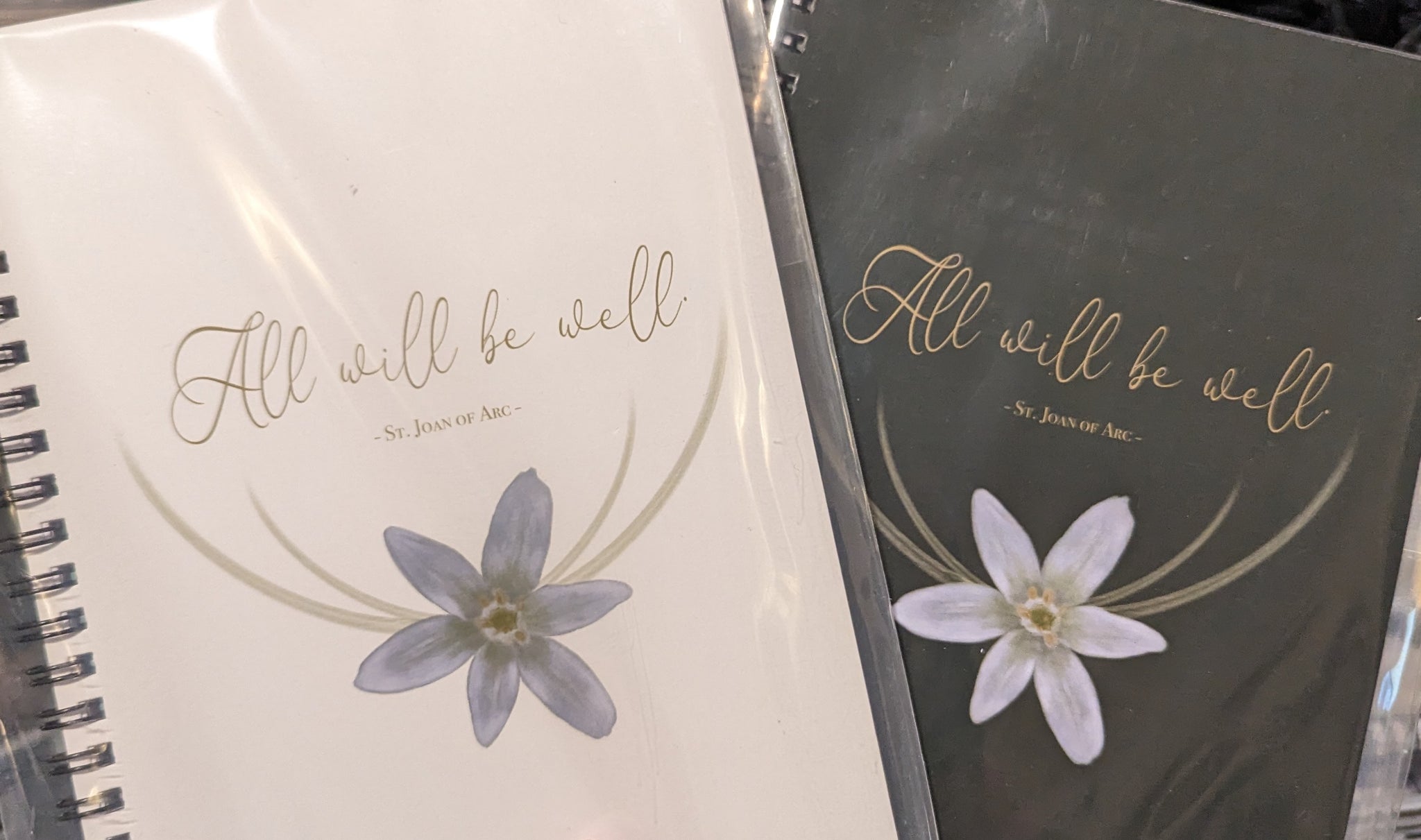 Two Catholic spiral notebooks or journals. The covers feature the quote from St. Joan of Arc "All will be Well" as well as the Star of Bethlehem flower.