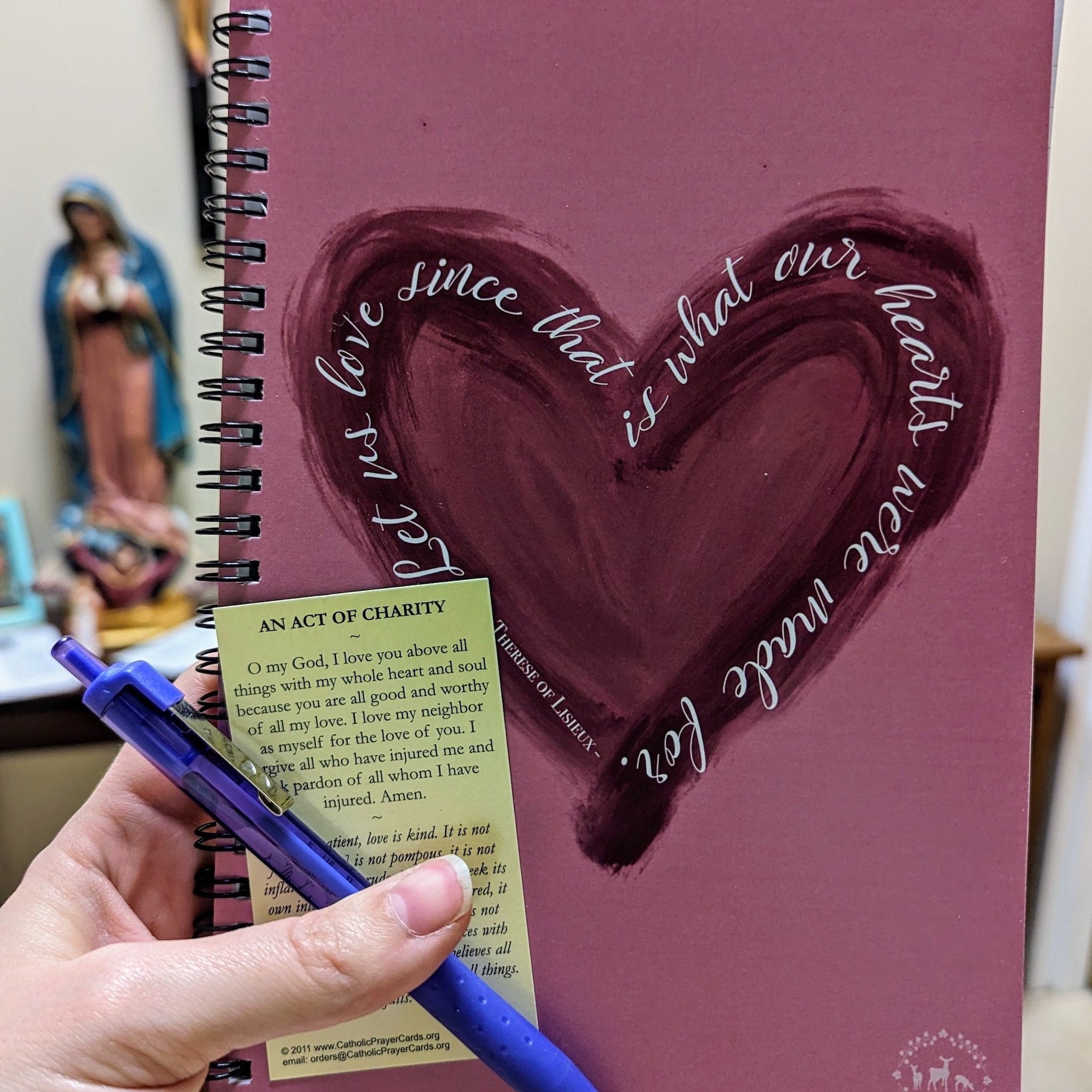 A Catholic notebook or journal with a heart on it and the St. Therese of Lisieux quote "Let us love since that is what our hearts were made for." The hand holding the notebook is also holding a pen and a Holy Card with the prayer "An Act of Charity" on it. 