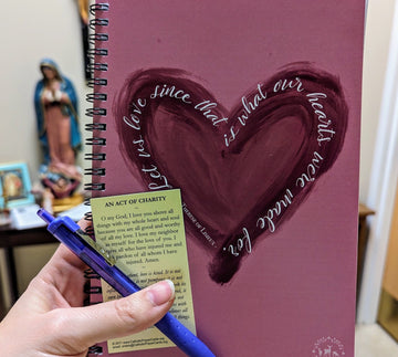 A Catholic notebook or journal with a heart on it and the St. Therese of Lisieux quote "Let us love since that is what our hearts were made for." The hand holding the notebook is also holding a pen and a Holy Card with the prayer "An Act of Charity" on it. 