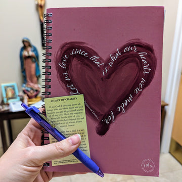 A Catholic notebook, or journal, with a heart on it and the St. Therese of Lisieux quote "Let us love since that is what our hearts were made for." The hand holding the notebook is also holding a pen and a Holy Card with the prayer "An Act of Charity" on it. 