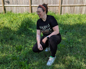 A woman kneels in the grass wearing a black Sacred Heart of Jesus shirt, glasses, a pink headband, and a smart watch.