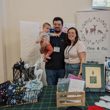 A man, woman, and baby behind the table of a vendor display featuring Catholic gift shop Ivy Doe & Co