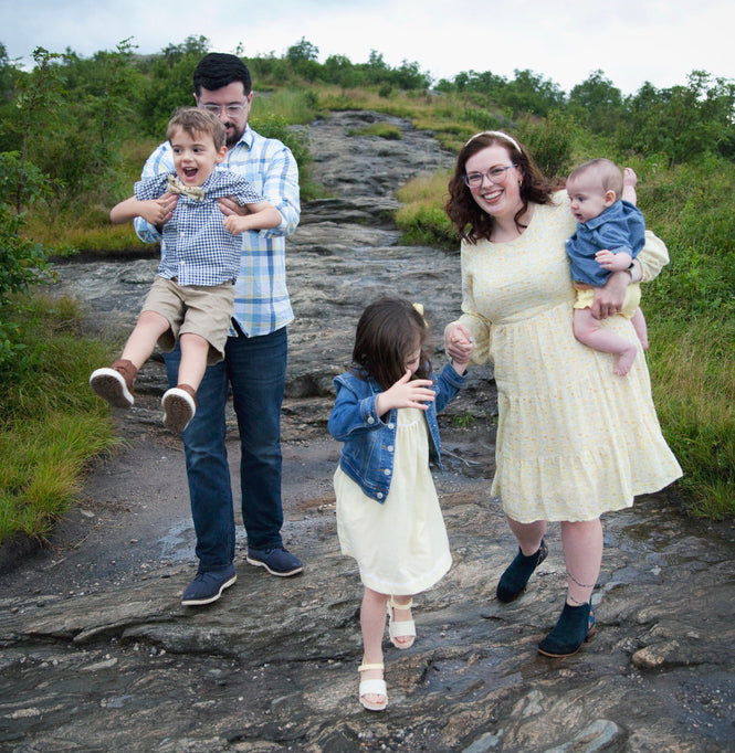 Photo of a Catholic family. A smiling mom holding a baby and a man or dad holding his son, while their daughter walks holding the woman's hand.